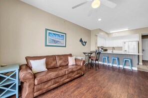 A living room with a brown leather sofa, a dining area with a small table and chairs, and a kitchen with white cabinets and a blue countertop. - Celadon 1505 - Turquoise Oasis (Panama City Beach)