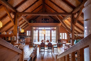 A rustic wooden interior with a high ceiling, a dining area with a round table, and large windows allowing natural light. - Exceptional Chalet - Panoramic View - Sauna - Aravis (Serraval)