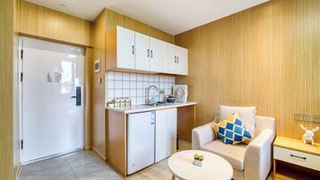 A compact kitchen with white cabinets, a small refrigerator, and a sink. There is a beige sofa with a patterned cushion and a small table with a plant.
