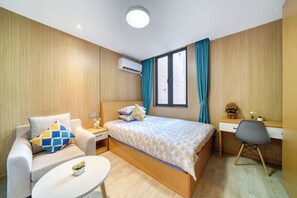 Quarto de hotel com cama, escrivaninha, cadeira, pequena mesa com planta e ar condicionado.