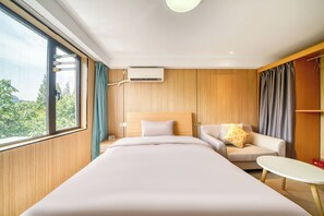A hotel room with a bed, a sofa, a small table, and a view of greenery outside.