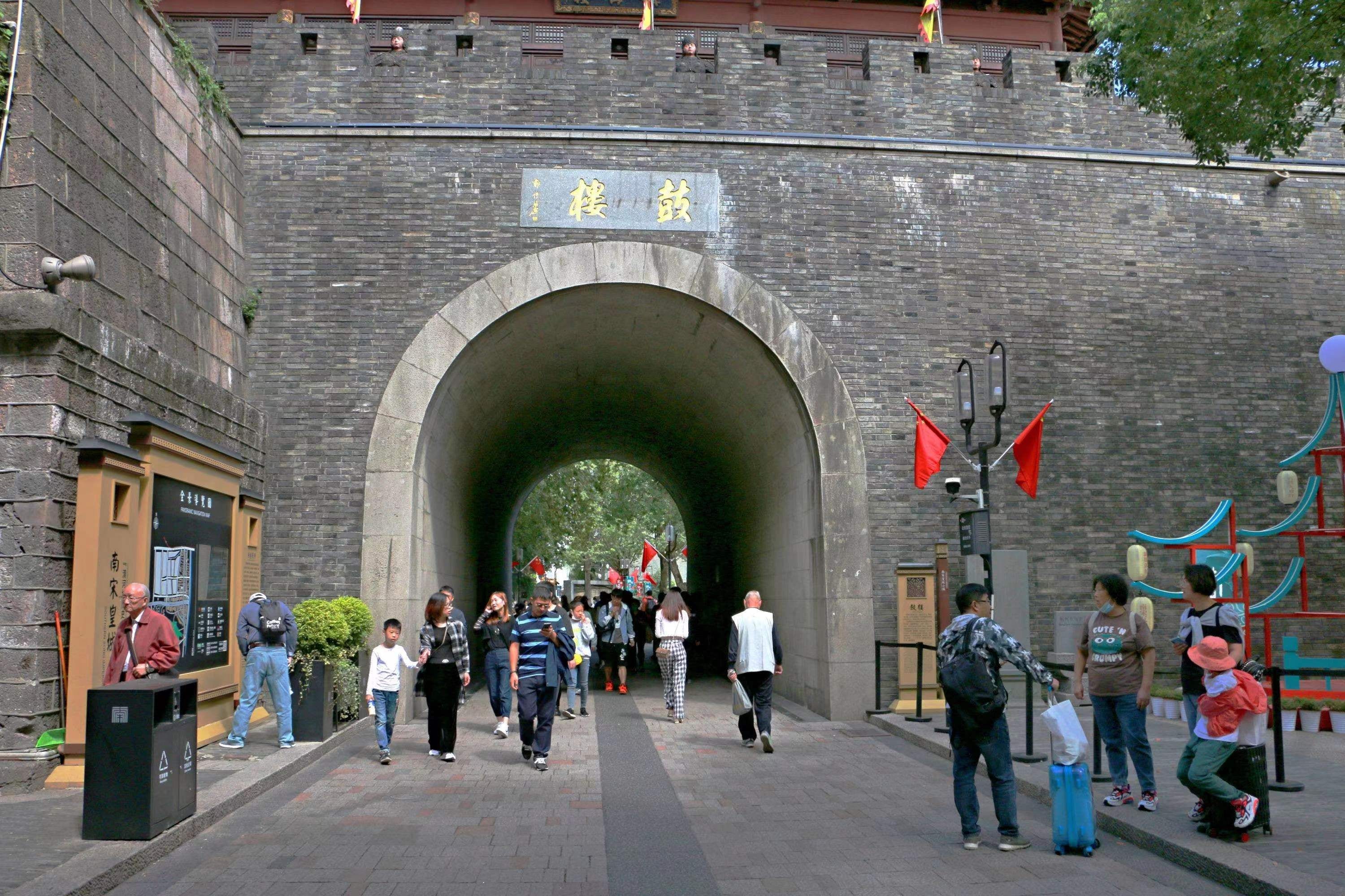 A stone archway leading into a tunnel with a sign above it. People are walking through the tunnel.