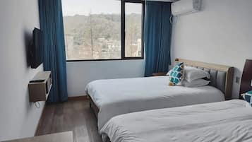 A hotel room with two beds, a TV, an air conditioning unit, and a window with a view of a cityscape.