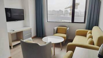 A modern living room with a flat-screen TV, a dining table, two sofas, and a large window with a view of a cityscape and construction cranes.