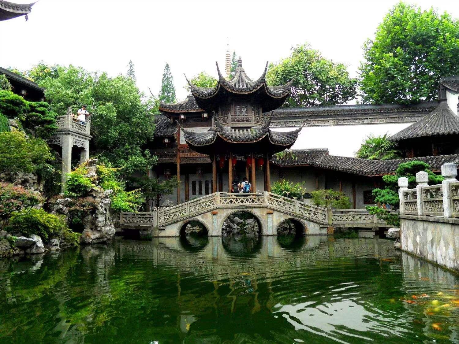 A traditional Chinese pavilion with a stone bridge over a pond, surrounded by lush greenery.