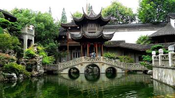 A traditional Chinese pavilion with a stone bridge over a pond, surrounded by lush greenery.