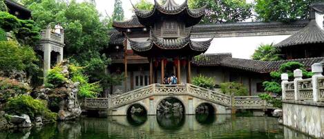 A traditional Chinese pavilion with a stone bridge over a pond, surrounded by lush greenery.