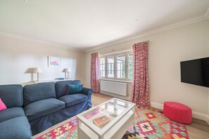 A living room with a sofa, a glass coffee table with a book, a red pouf, and a window with patterned curtains. - Summerfield House, Blandford Forum, Wimborne (Blandford Forum)
