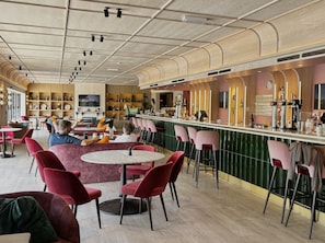 Ein stilvoller Barbereich mit runden Tischen und roten Stühlen, eine Theke mit Barbekleidung und Regale mit verschiedenen Gegenständen.