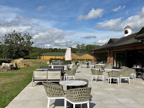 Un espace extérieur aménagé en terrasse, avec des tables rondes et des chaises en osier, un grand parasol, et un bâtiment avec un clocher en arrière-plan.