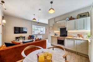 A modern kitchen with a dining area, featuring a flat-screen TV, pendant lights, and a breakfast setup with orange juice. - Foxglove Cottage, Hamptworth, Hampshire (Salisbury)