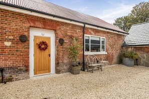 A brick building with a wooden door, a window, a bench, and a wreath. - Foxglove Cottage, Hamptworth, Hampshire (Salisbury)