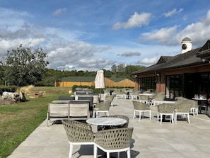 An outdoor patio area with round tables and wicker chairs, a large umbrella, and a building with a clock tower in the background. - Fern Cottage, Hamptworth, Hampshire (Salisbury)