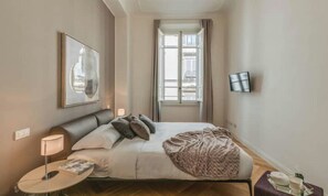 Une chambre d’hôtel avec un lit, une table de chevet avec une lampe, un tableau encadré au mur et une fenêtre avec des rideaux.