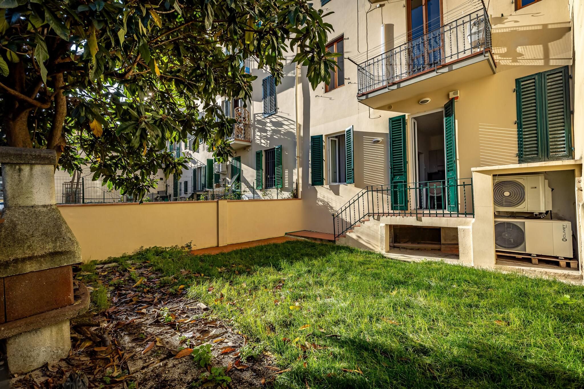 A courtyard with a stone bench, a tree, and a building with green shutters and a balcony.