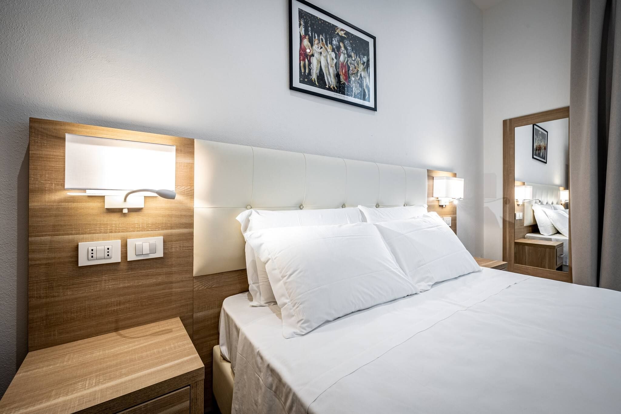 A hotel room with a wooden headboard, a large bed with white linens, bedside lamps, a nightstand, and a framed picture on the wall.