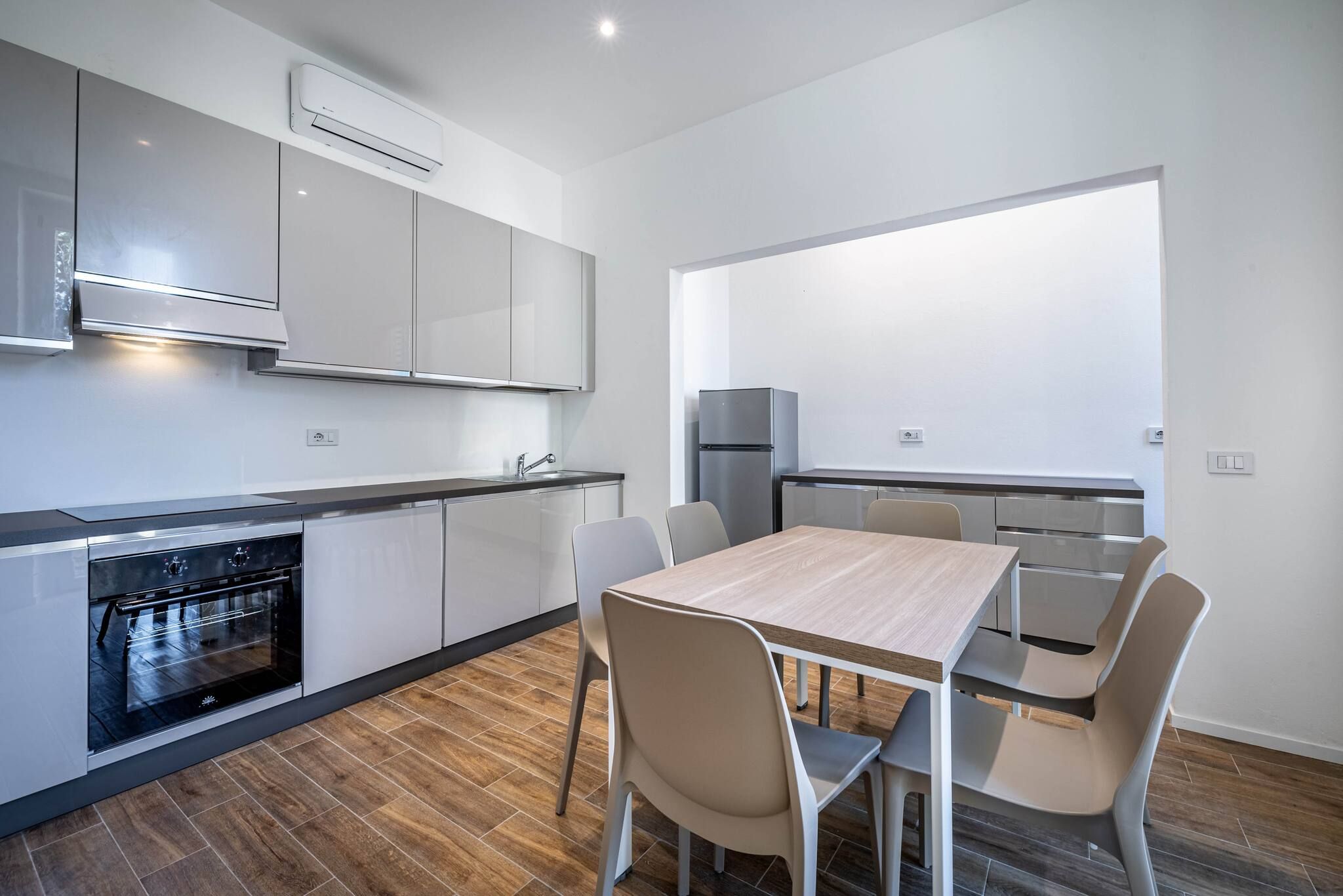 Una cocina moderna con armarios blancos, horno empotrado y comedor con mesa y sillas de madera.