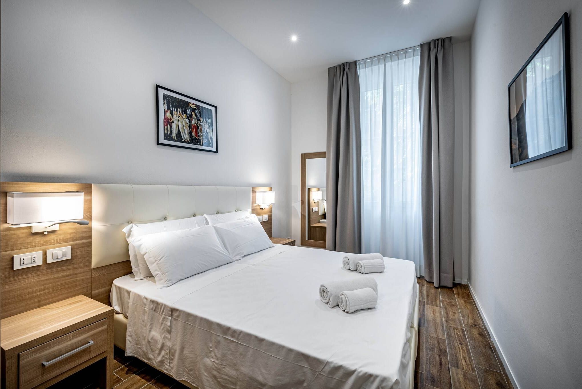 Une chambre d’hôtel avec un grand lit, des lampes de chevet, une table de chevet, une tête de lit en bois, un tableau encadré au mur et une fenêtre avec des rideaux.