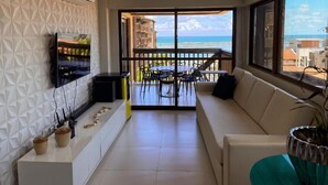 A modern living room with a balcony overlooking a beach, a flat-screen TV mounted on the wall, and a dining area with chairs and a table. - Cobertura Vista Mar Barra Bali (Barra de São Miguel)