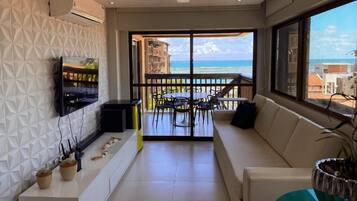Ein modernes Wohnzimmer mit Balkon und Meerblick, einem Wand-Fernseher und einem Essbereich mit Stühlen und Tisch.