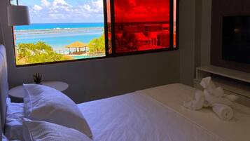 Quarto de hotel com cama, televisão, janela com vista para a praia e uma pequena mesa com uma planta.
