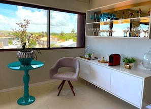 Quarto com vista para a paisagem urbana através de amplas janelas, uma mesa verde-água com planta em vaso, um armário branco com copos e uma cadeira rosa.