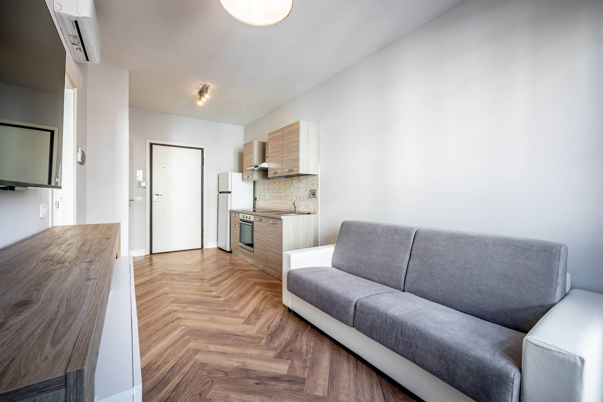A modern living room with a grey sofa, wooden flooring, a kitchen with light wood cabinets, and a flat-screen TV mounted on the wall.