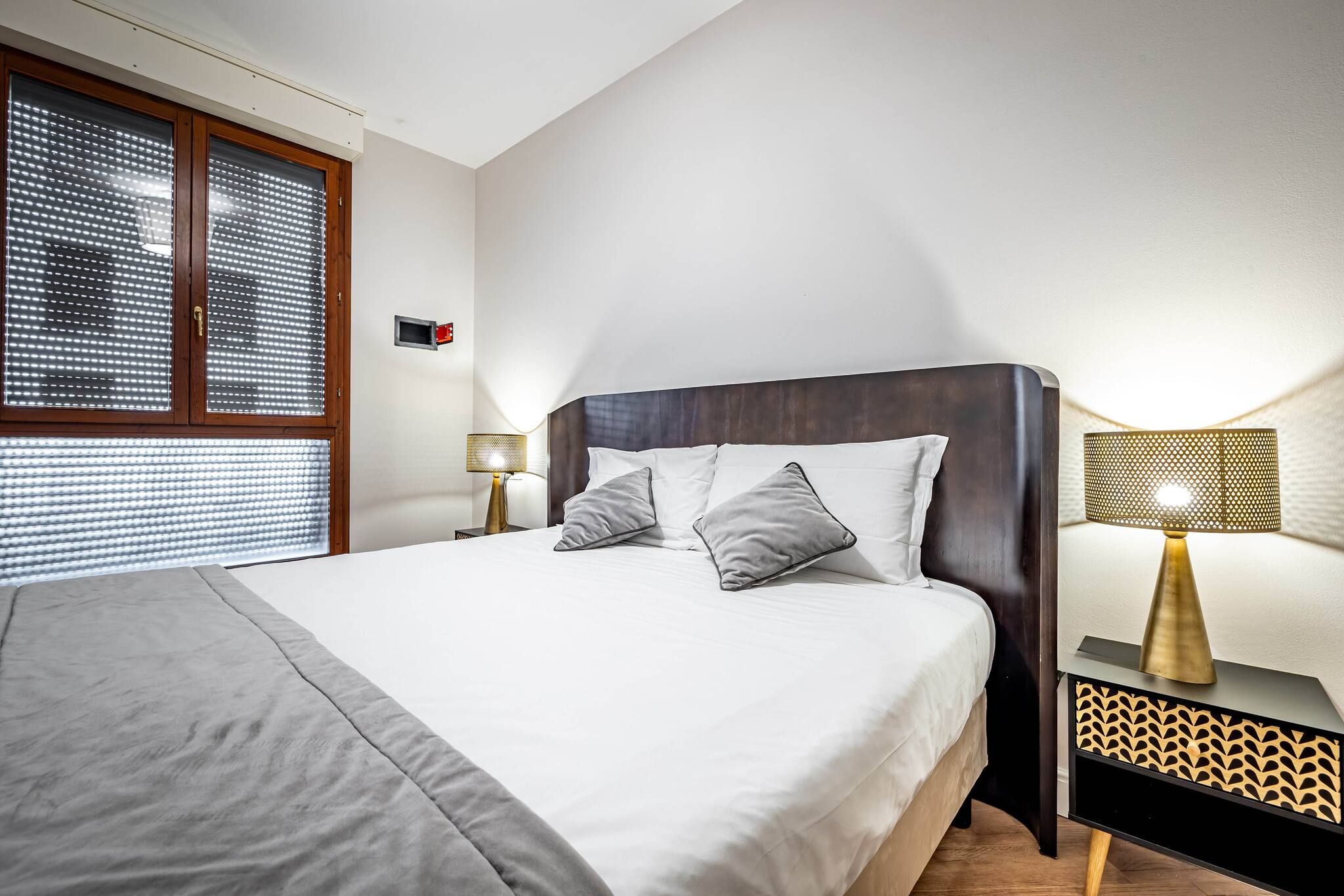 A neatly made bed with a grey blanket, two pillows, and a wooden headboard. There is a bedside table with a lamp and a wall-mounted alarm clock.