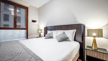 A neatly made bed with a grey blanket, two pillows, and a wooden headboard. There is a bedside table with a lamp and a wall-mounted alarm clock.