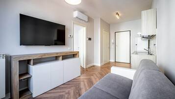 Una sala de estar moderna con un televisor de pantalla plana, almacenamiento integrado, un sofá gris y una cocina con estufa y armarios.