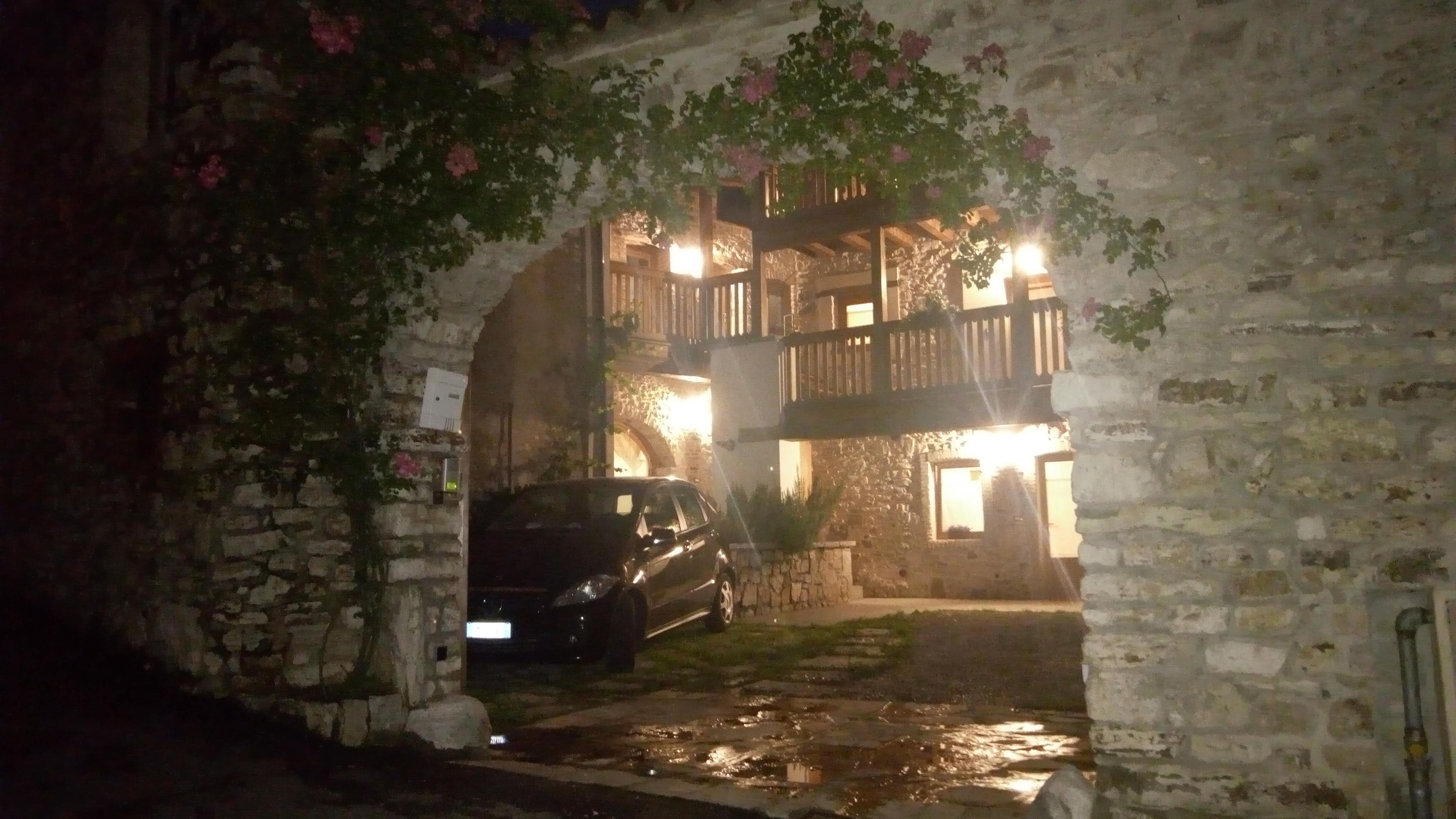 A stone building with a car parked in front, a balcony, and a small garden area.
