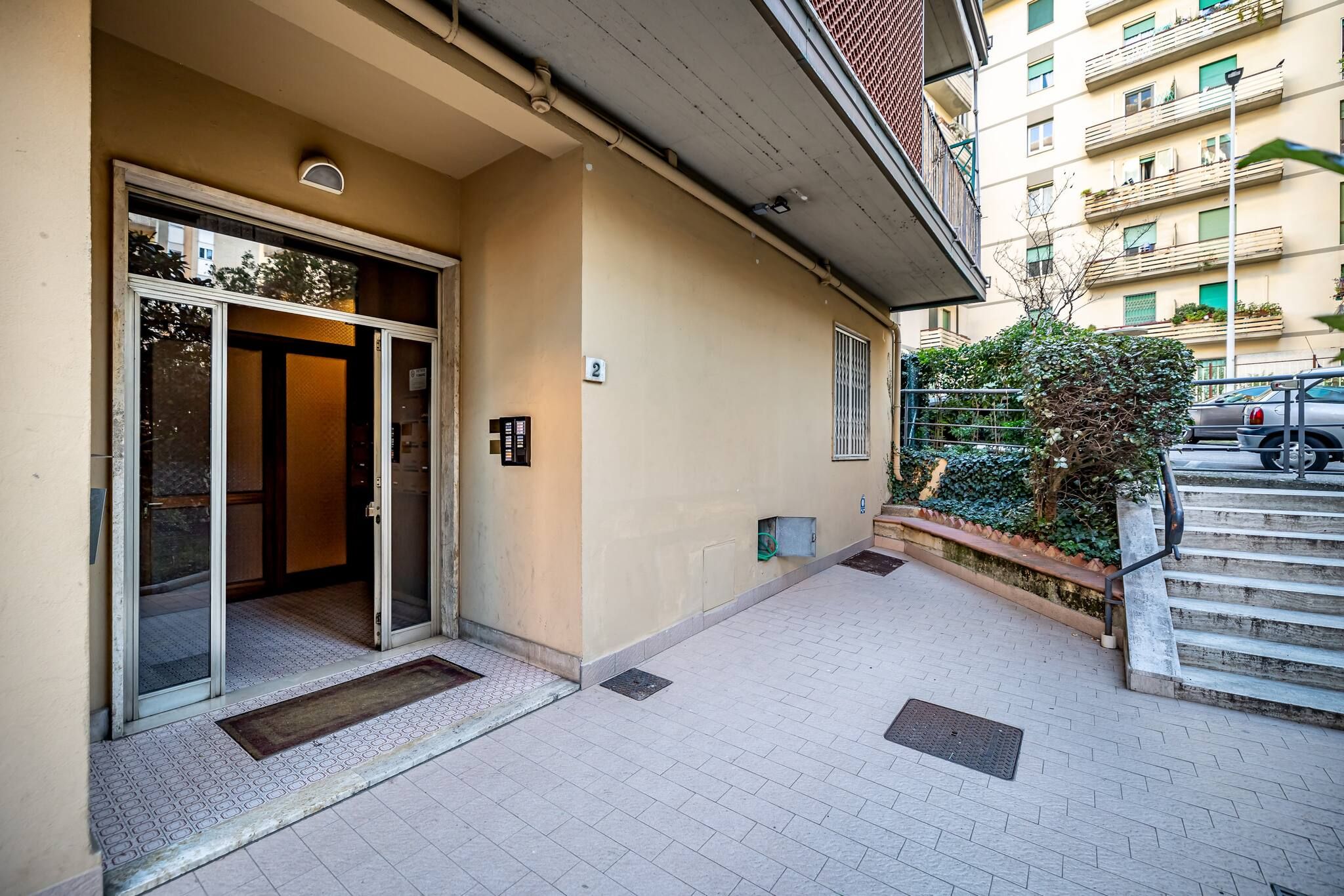 A building entrance with a glass door, a number plaque, a small garden area, and a staircase leading to an upper level.