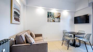 A modern living room with a sofa, a dining table with chairs, a flat-screen TV mounted on the wall, and artwork on the walls.