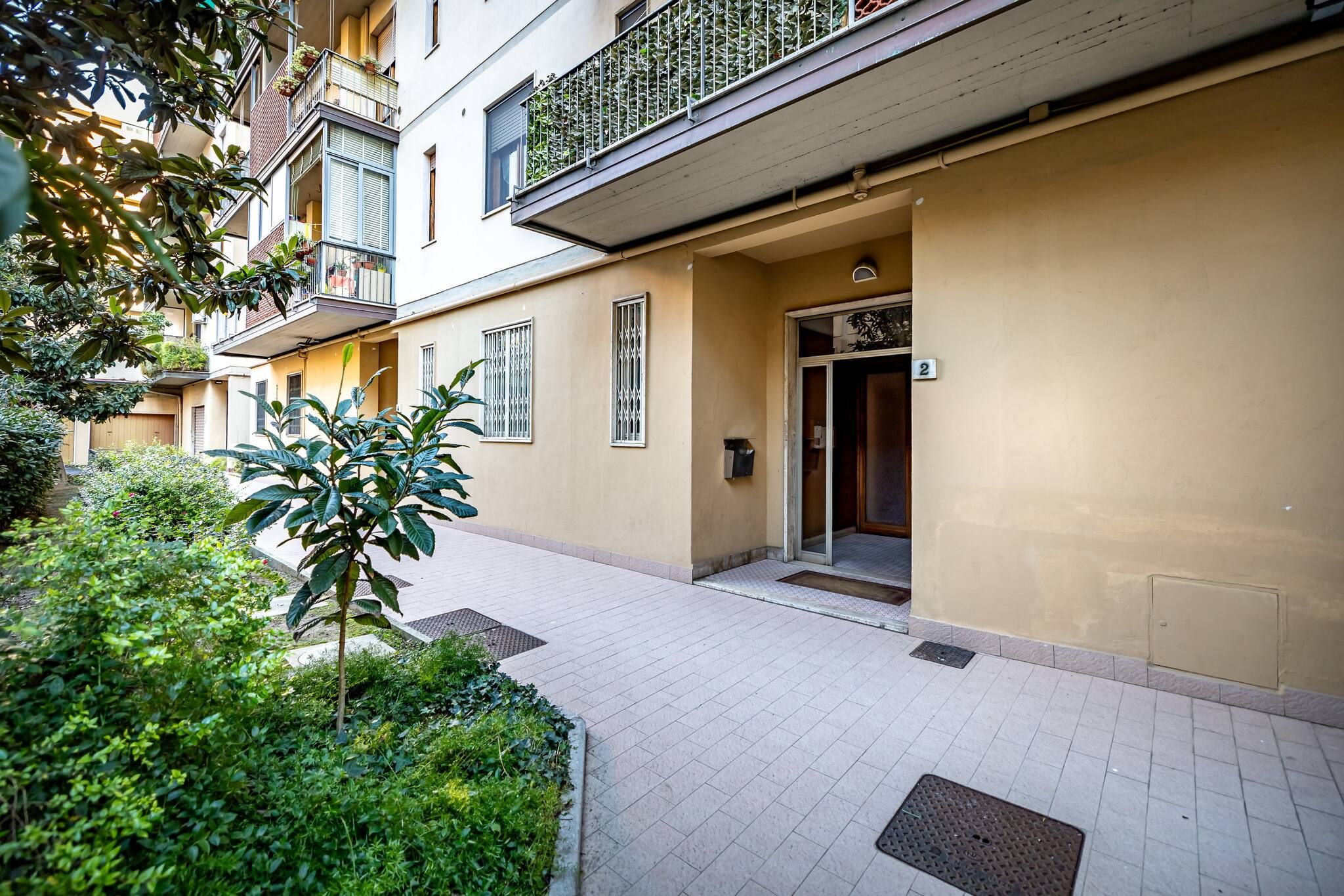 A multi-story residential building with a tiled entrance, a small garden area, and balconies with plants.
