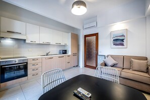 Et moderne køkken med hvide skabe, en sort bordplade og en indbygget ovn. Der er et spisebord med stole og en fjernbetjening på. En brun sofa står op ad væggen med et indrammet abstrakt billede over.