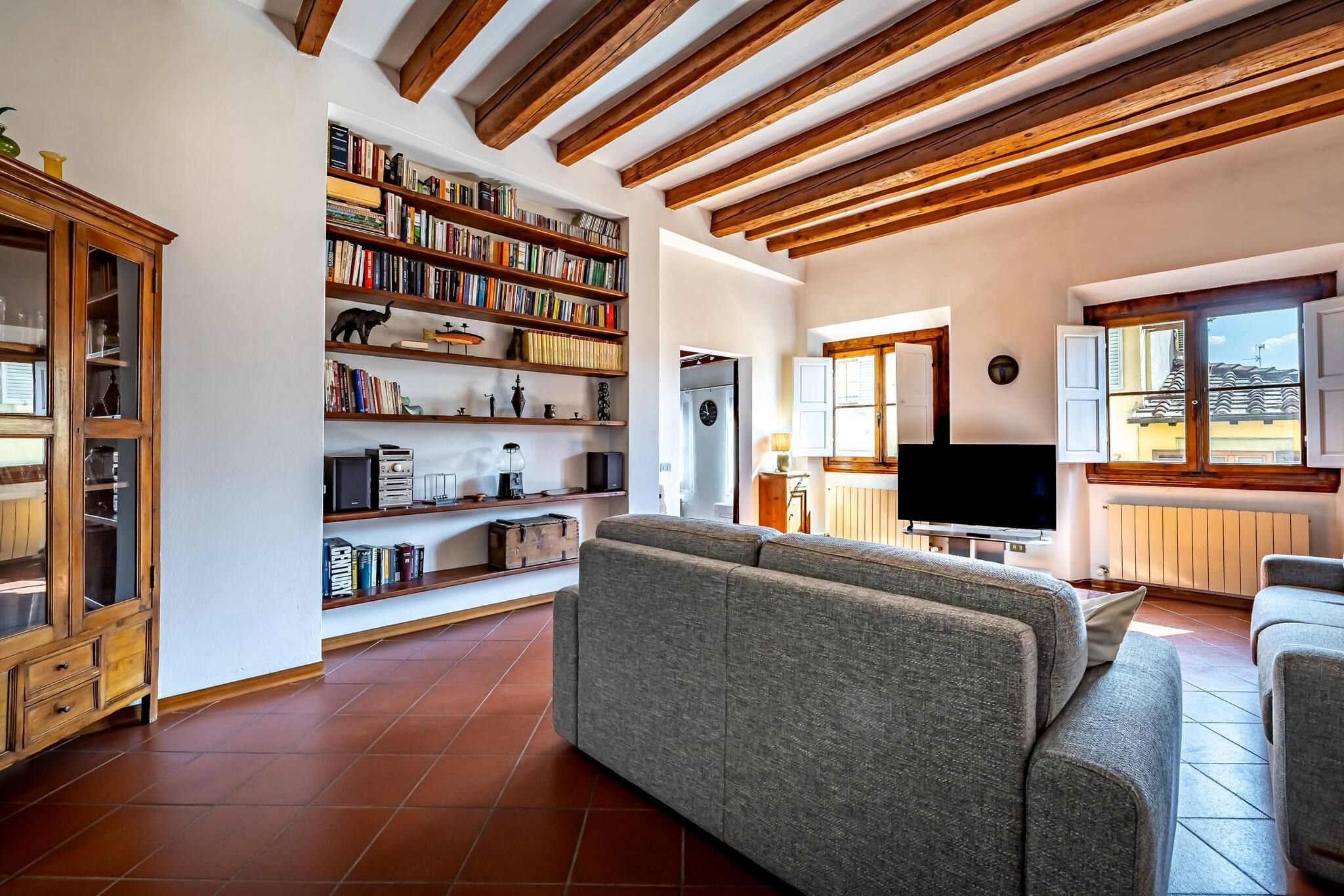 Ein Wohnzimmer mit grauem Sofa, Holzbücherregalen, einem Fernseher und einer durchgehenden Tür erkennbaren Küchenzeile.