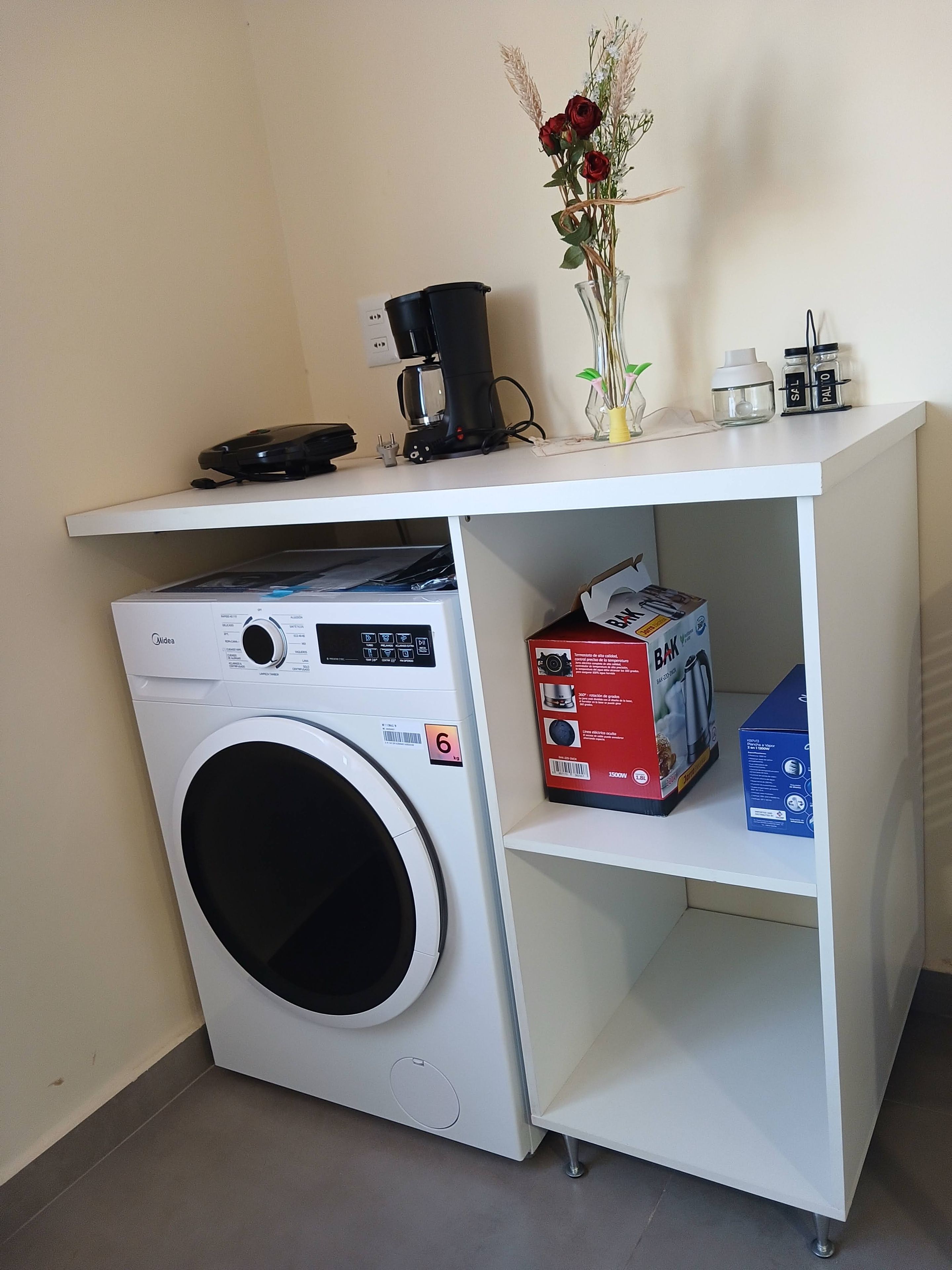 A compact laundry area with a front-loading washing machine, a coffee maker, and a small shelf with a box of BAK detergent.