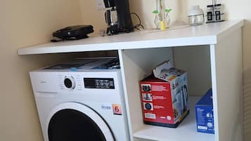 A compact laundry area with a front-loading washing machine, a coffee maker, and a small shelf with a box of BAK detergent.