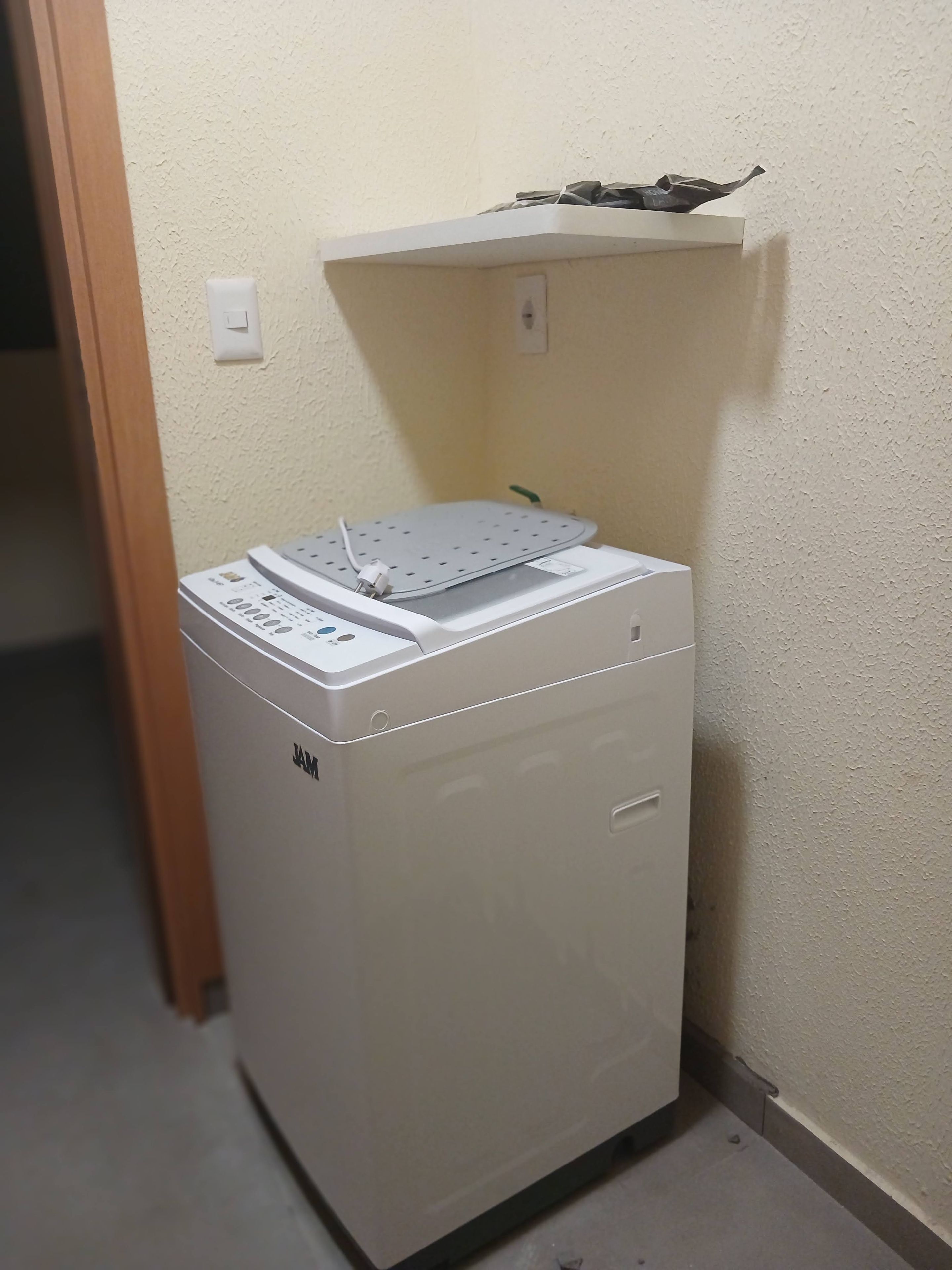 A compact, white, front-loading washing machine with a control panel and a drying rack on top, situated in a corner with a textured wall and a wooden door frame.