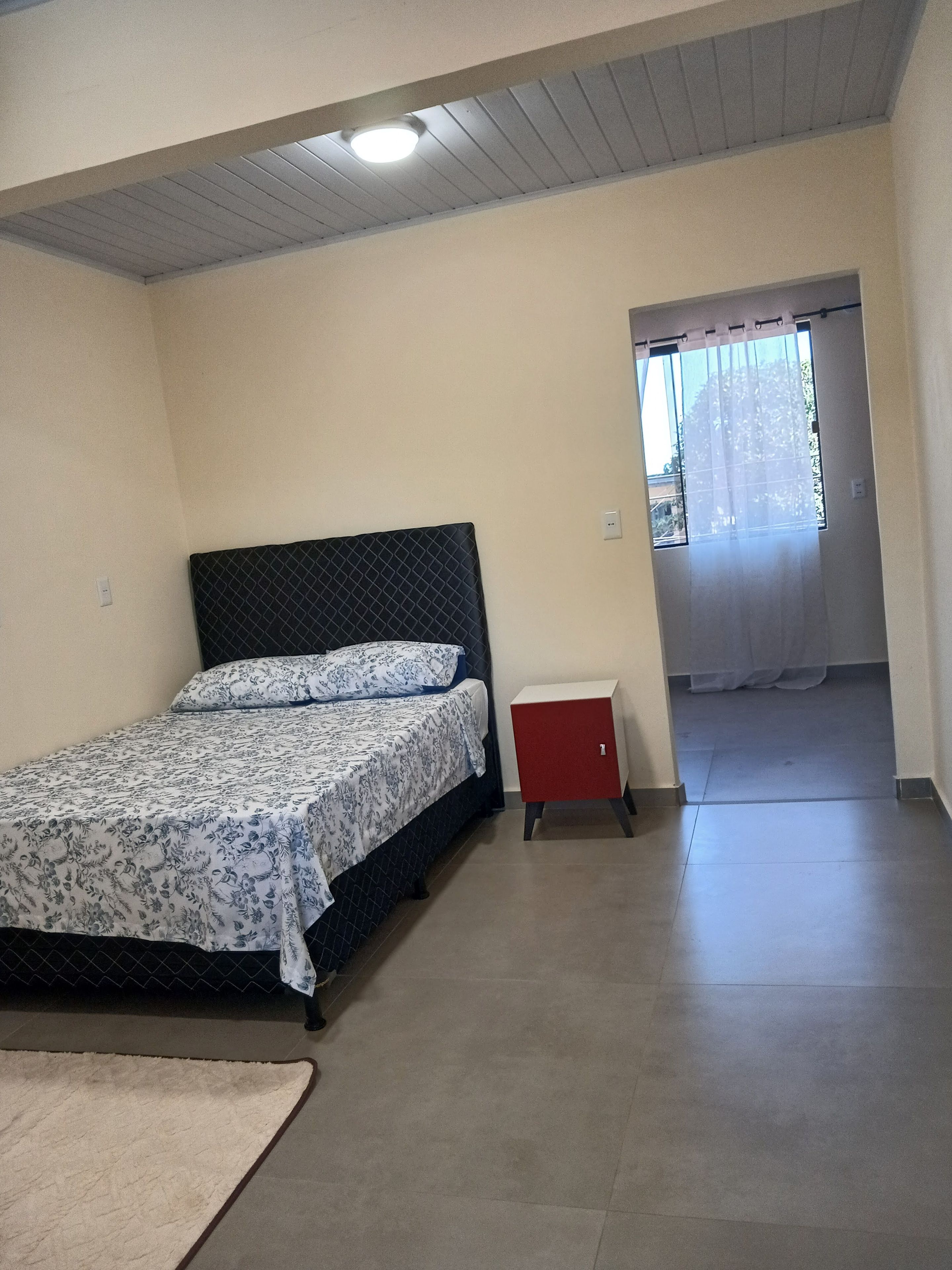 Ein Schlafzimmer mit einem Bett, einem kleinen roten Schrank und einem Fenster mit Vorhängen.