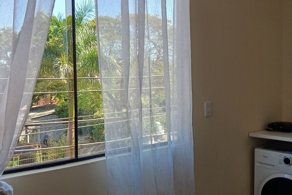 A room with a window covered by sheer curtains, a washing machine, and a metal railing outside.