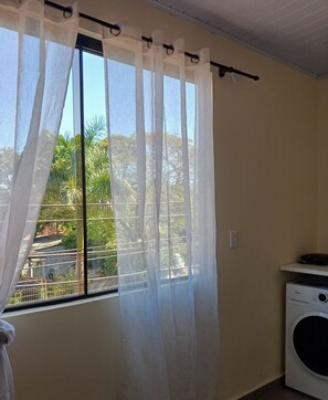 Habitación con ventana cubierta por cortinas de tul, lavadora y baranda de metal en el exterior.
