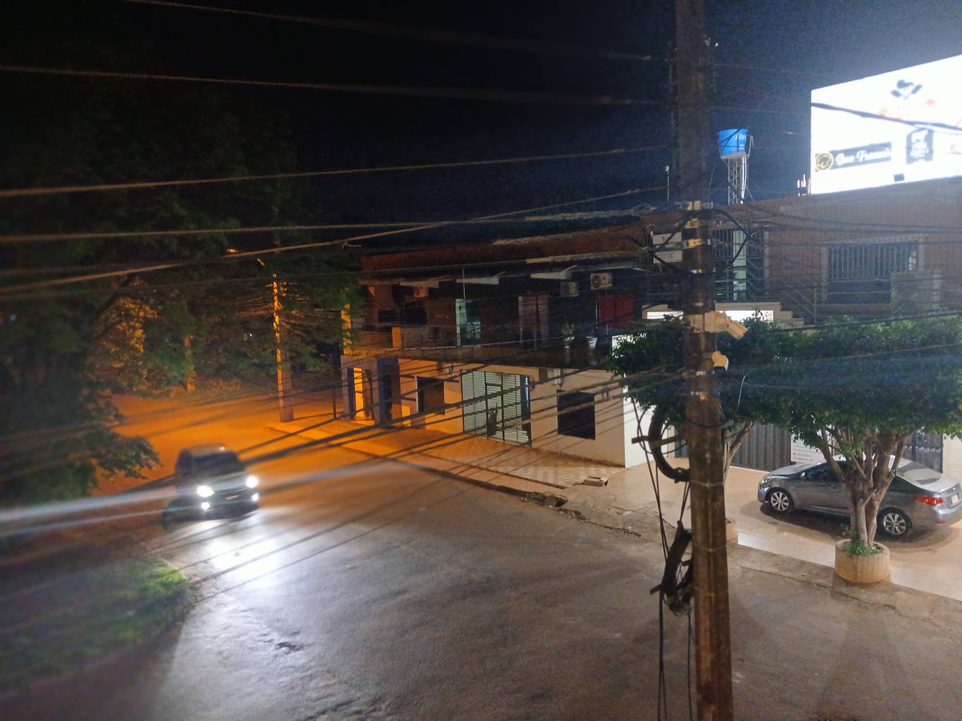 Vista noturna da rua com um prédio, um outdoor e carros estacionados.