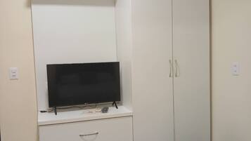 Ein Zimmer mit einem weißen Kleiderschrank, einem Flachbildfernseher auf einem Standfuß, einem kleinen Hocker und einer an der Wand montierten Klimaanlage.