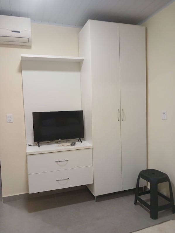 A room with a white wardrobe, a flat-screen TV on a stand, a small stool, and an air conditioner mounted on the wall.