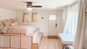 A bedroom with a bed, a bedside table, a chair, a window with curtains, a ceiling fan, and a door.