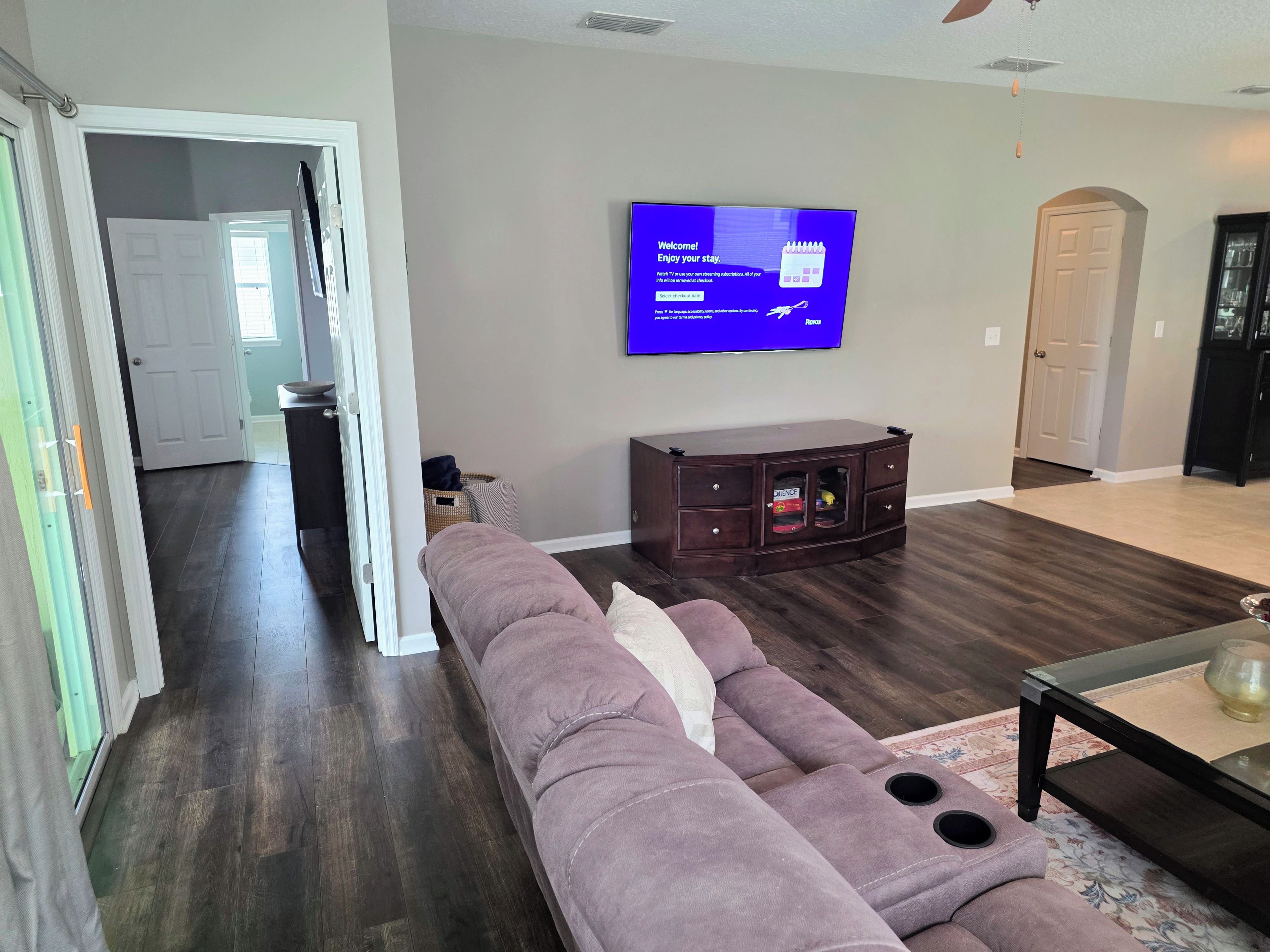 A living room with a sectional sofa, a flat-screen TV mounted on the wall, a dark wood cabinet, and a glass coffee table.