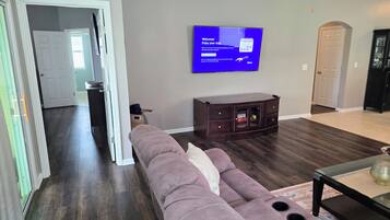 A living room with a sectional sofa, a flat-screen TV mounted on the wall, a dark wood cabinet, and a glass coffee table.