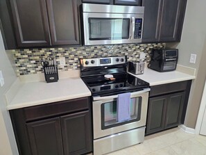 A kitchen with dark cabinets, a stainless steel microwave, a stove, and a toaster. - Modern Open Concept 3/2 Retreat (Orange Park)