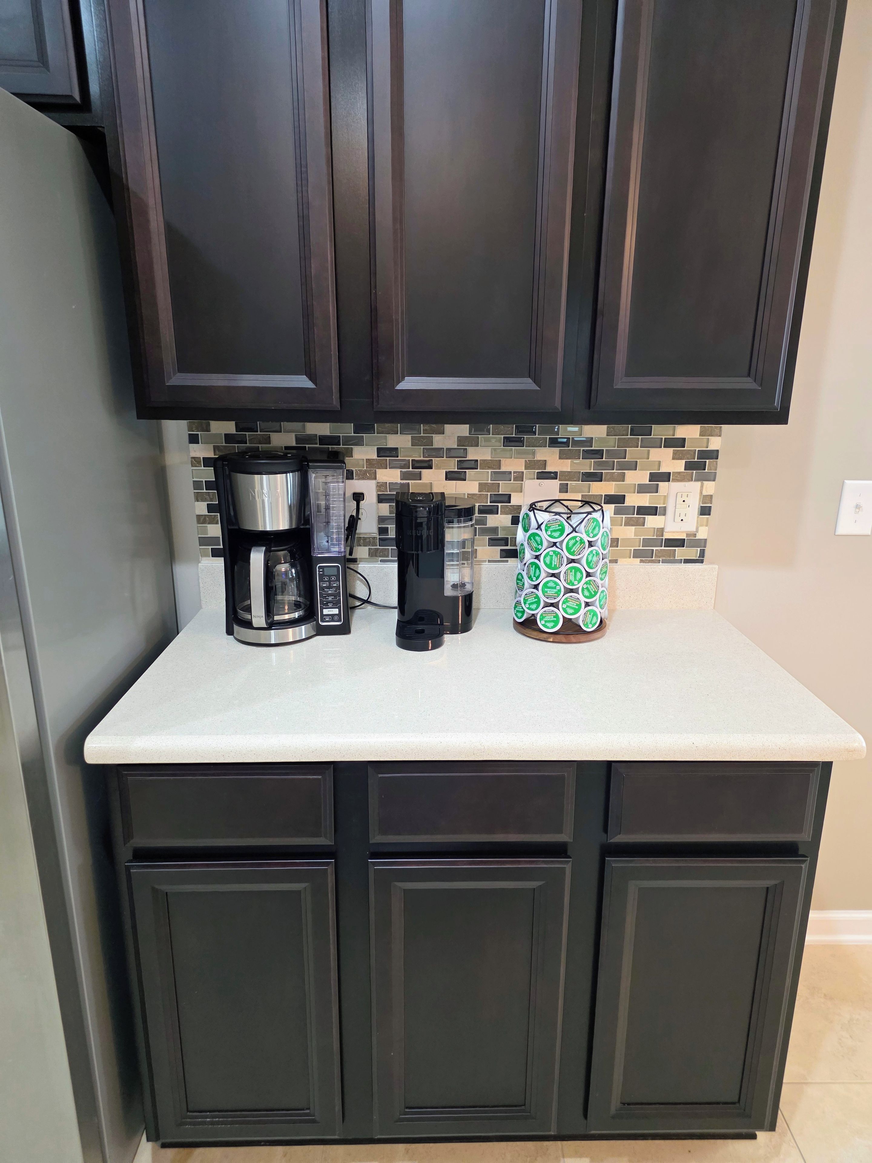 A kitchen with dark cabinets, a coffee maker, a blender, and a coffee pod holder.